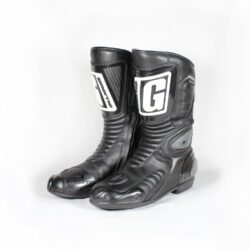 A pair of used black Gaerne G-RT racing motorcycle boots in size 43, made of leather. Features include heel, toe, gear lever, ankle, and shin protection, a non-slip oil-resistant sole, a wrap-around heel cup, and a double closure system with a zip and wide Velcro strap. Shows scratching on the plastic protectors.