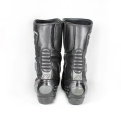 A pair of used black Gaerne G-RT racing motorcycle boots in size 43, made of leather. Features include heel, toe, gear lever, ankle, and shin protection, a non-slip oil-resistant sole, a wrap-around heel cup, and a double closure system with a zip and wide Velcro strap. Shows scratching on the plastic protectors.