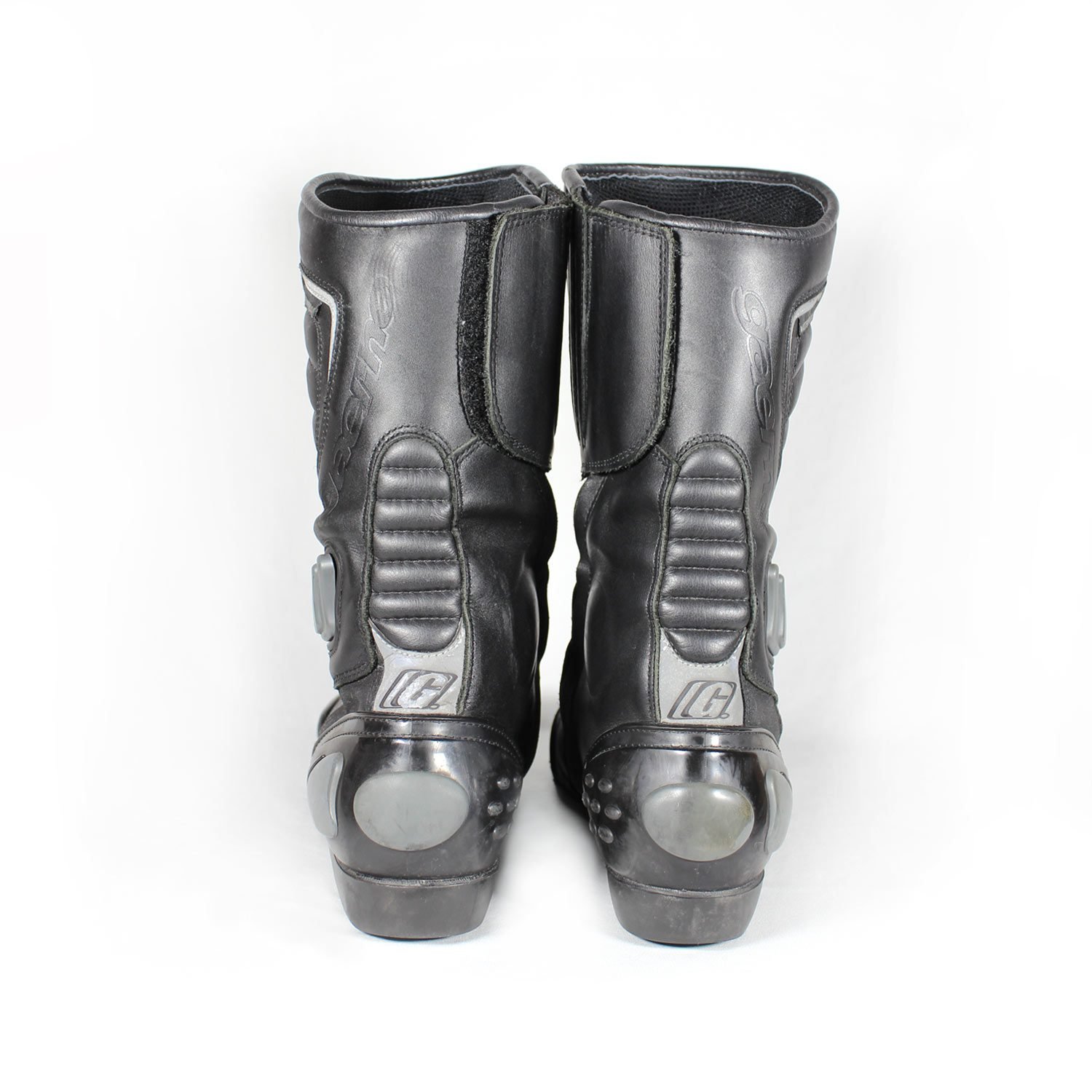 A pair of used black Gaerne G-RT racing motorcycle boots in size 43, made of leather. Features include heel, toe, gear lever, ankle, and shin protection, a non-slip oil-resistant sole, a wrap-around heel cup, and a double closure system with a zip and wide Velcro strap. Shows scratching on the plastic protectors.