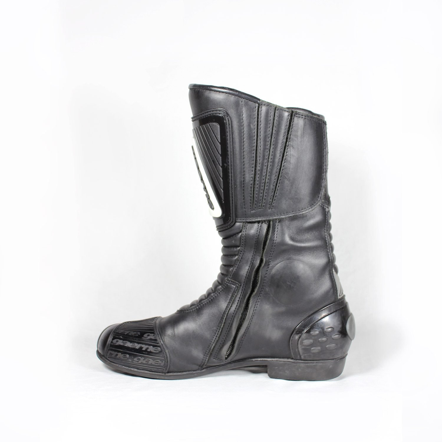 A pair of used black Gaerne G-RT racing motorcycle boots in size 43, made of leather. Features include heel, toe, gear lever, ankle, and shin protection, a non-slip oil-resistant sole, a wrap-around heel cup, and a double closure system with a zip and wide Velcro strap. Shows scratching on the plastic protectors.