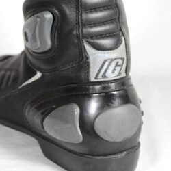 A pair of used black Gaerne G-RT racing motorcycle boots in size 43, made of leather. Features include heel, toe, gear lever, ankle, and shin protection, a non-slip oil-resistant sole, a wrap-around heel cup, and a double closure system with a zip and wide Velcro strap. Shows scratching on the plastic protectors.