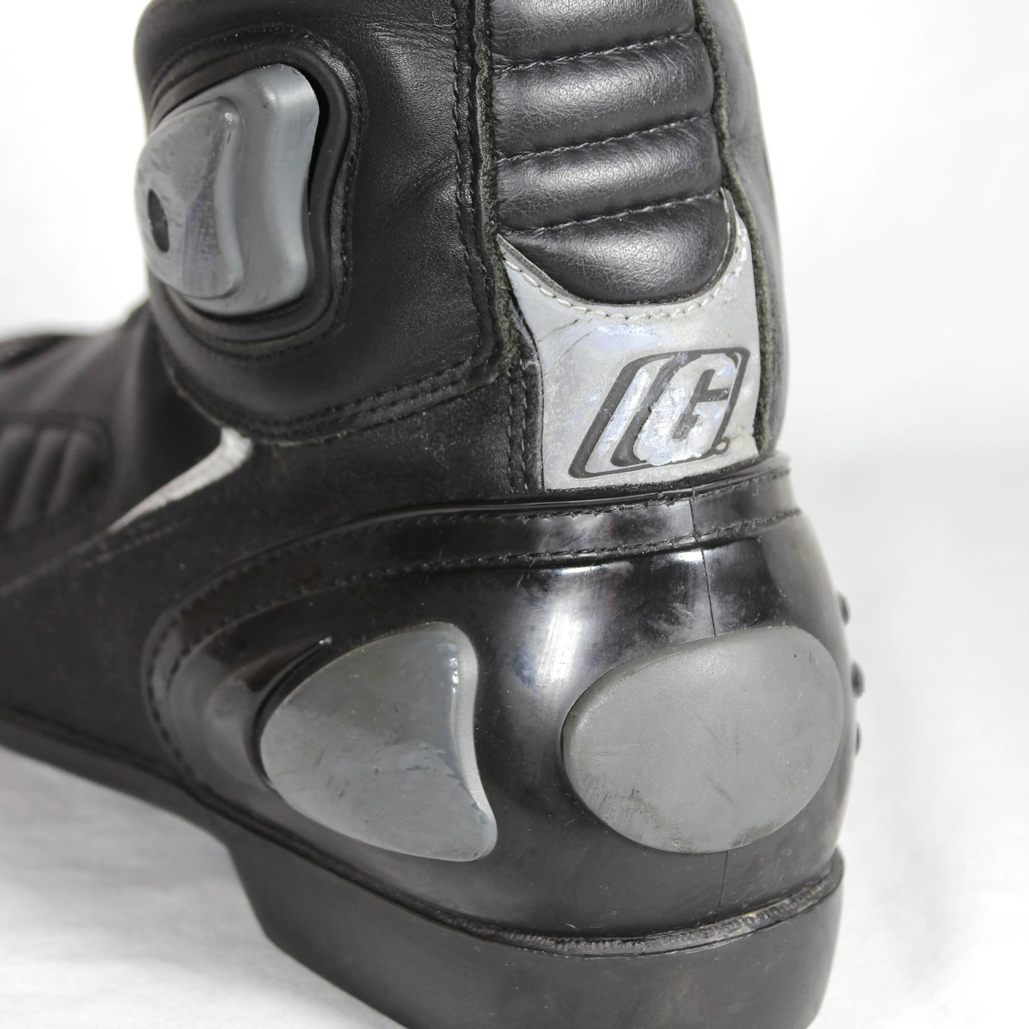 A pair of used black Gaerne G-RT racing motorcycle boots in size 43, made of leather. Features include heel, toe, gear lever, ankle, and shin protection, a non-slip oil-resistant sole, a wrap-around heel cup, and a double closure system with a zip and wide Velcro strap. Shows scratching on the plastic protectors.