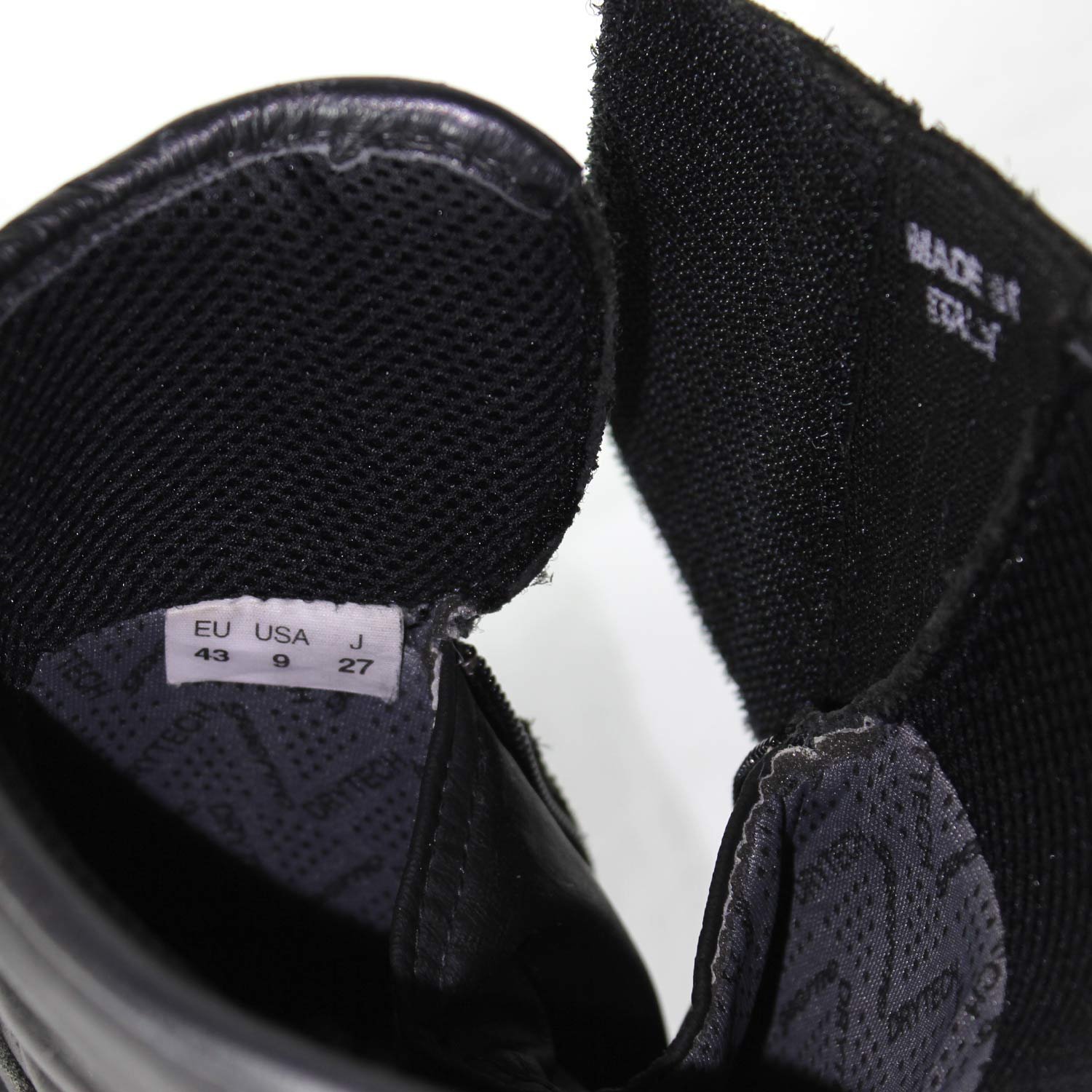 A pair of used black Gaerne G-RT racing motorcycle boots in size 43, made of leather. Features include heel, toe, gear lever, ankle, and shin protection, a non-slip oil-resistant sole, a wrap-around heel cup, and a double closure system with a zip and wide Velcro strap. Shows scratching on the plastic protectors.