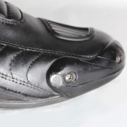 A pair of used black Gaerne G-RT racing motorcycle boots in size 43, made of leather. Features include heel, toe, gear lever, ankle, and shin protection, a non-slip oil-resistant sole, a wrap-around heel cup, and a double closure system with a zip and wide Velcro strap. Shows scratching on the plastic protectors.