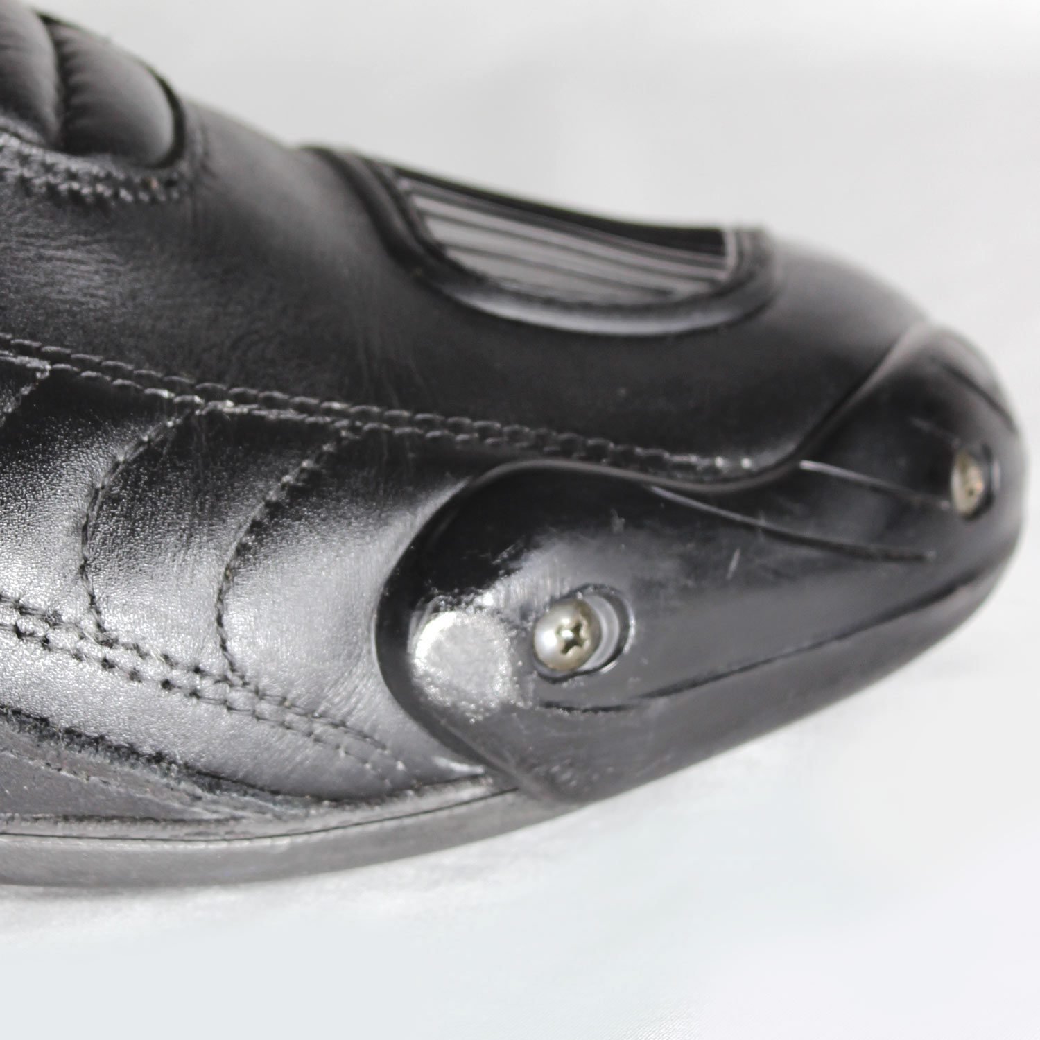 A pair of used black Gaerne G-RT racing motorcycle boots in size 43, made of leather. Features include heel, toe, gear lever, ankle, and shin protection, a non-slip oil-resistant sole, a wrap-around heel cup, and a double closure system with a zip and wide Velcro strap. Shows scratching on the plastic protectors.